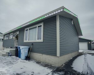 A gray house with green holiday lights under a cloudy sky, surrounded by snow-covered ground.