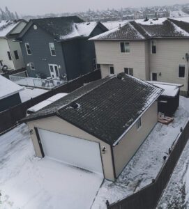 Aerial view of snow-covered residential houses with dark rooftops and a fenced yard.